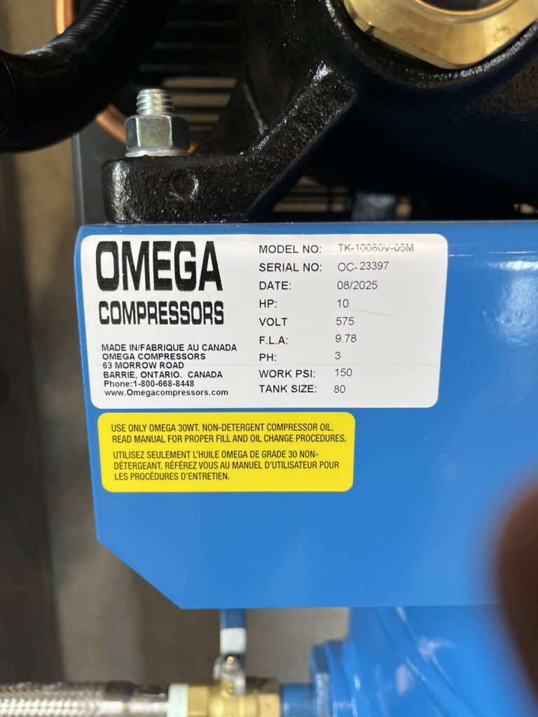 A blue Omega compressor, positioned on-site, is shown with its exterior blue casing, specification labels, and one visible pipe connection attached to the unit.